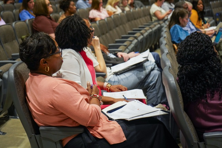 Women sit in chairs in an auditorium and take notes as a speaker talks  