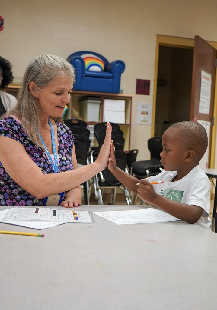 A teacher smiles and gives a child a high five while they sit at a table inside a classroom.