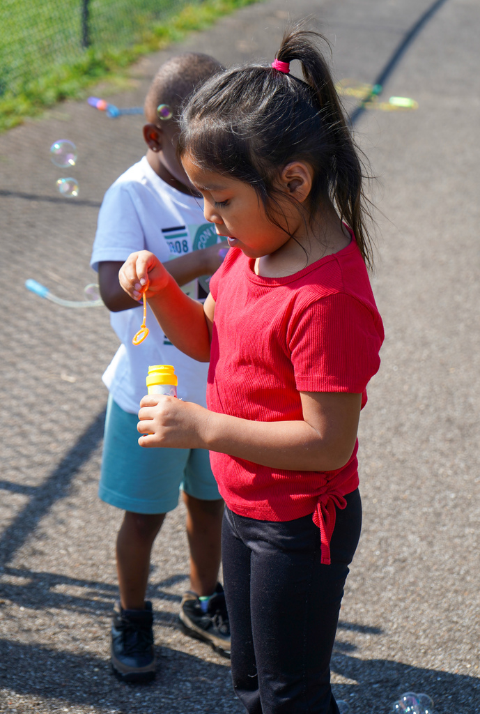 A girl blows bubbles while standing on a sidewalk outside.
