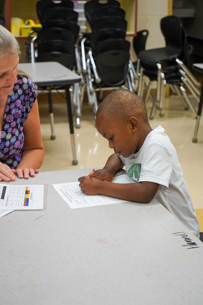 A boy writes on a piece of paper while sitting at a table with a teacher inside a classroom.
