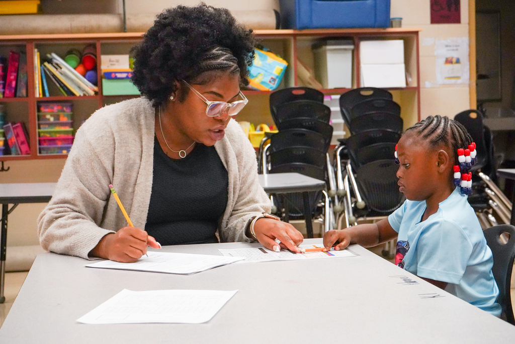 A teacher speaks to a child and points at a piece of paper while they sit at a table inside a classroom.