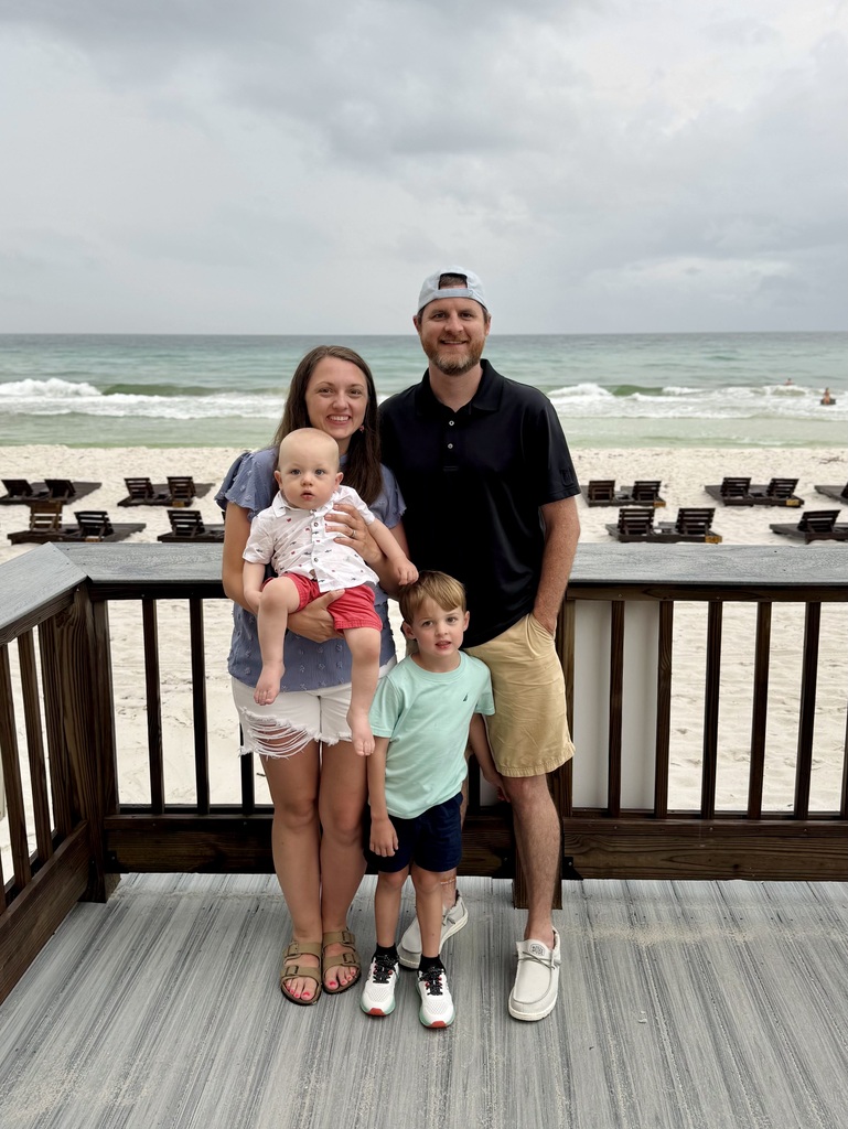 A man and a woman stand together with their children for a photo on a deck in front of a beach.