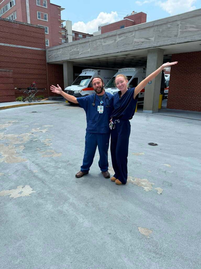 A man and a woman stand together for a photo outside a hospital. They smile and wear scrubs.