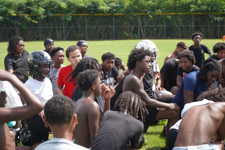 Football players sit on a grassy field and listen to their coach.
