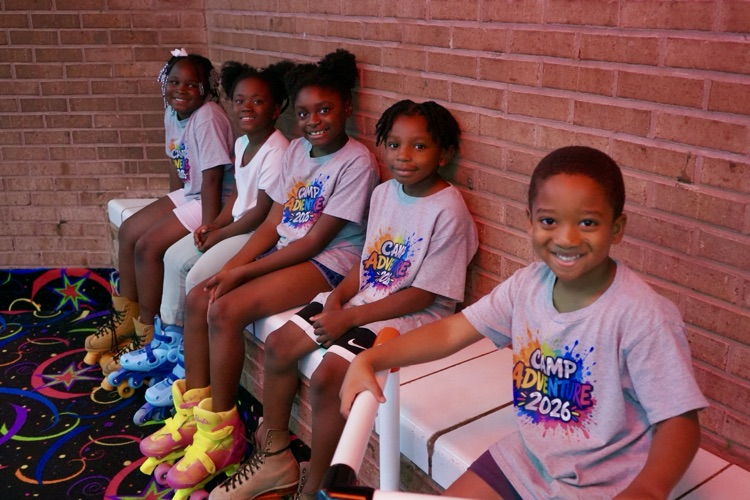 A group of children sit on a bench at a skating rink and smile for a photo