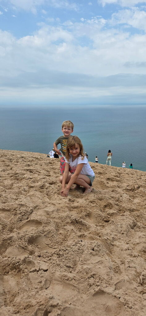 A boy and a girl smile for a photo at Sleeping Bear Dunes. They stand on the sand overlooking the water far below.