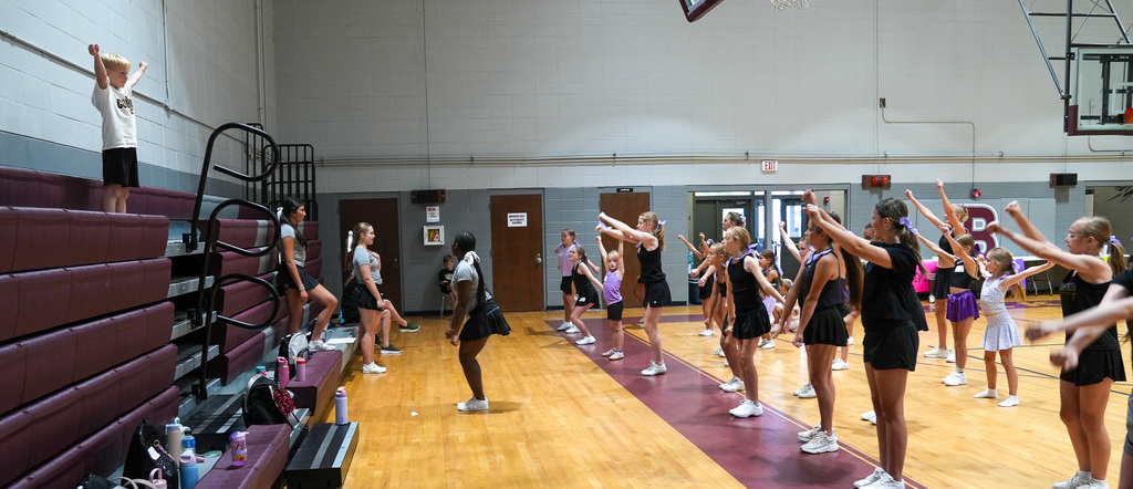 Cheerleaders hold their arms in the air during a cheer clinic at a school gym.