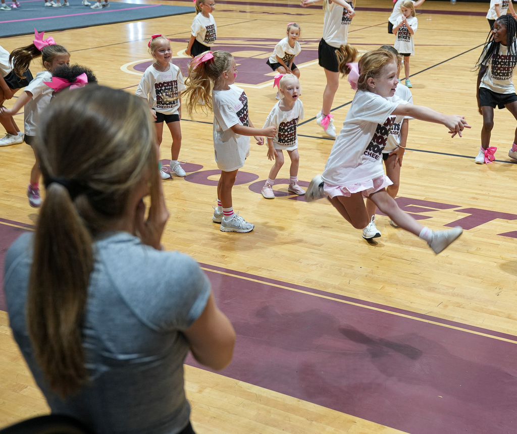 A young cheerleader jumps in the air during a cheer clinic inside a school gym.