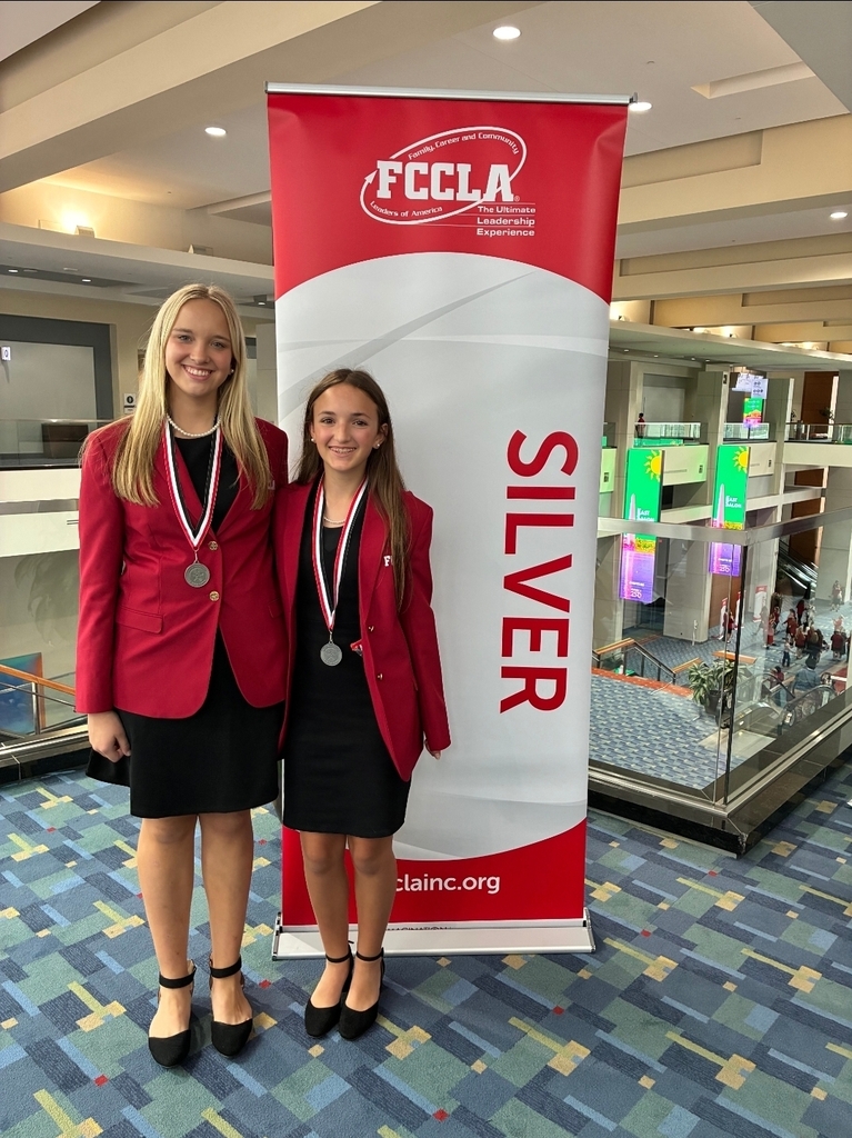 Two girls stand in professional clothing wearing medals around their necks standing in front of a banner that says silver