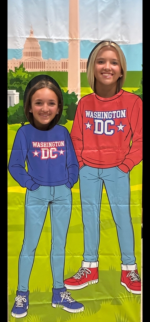 two girls stand behind a cut out with their faces showing through two holes. The front shows two cartoon like bodies in sweatshirts that say Washignton DC