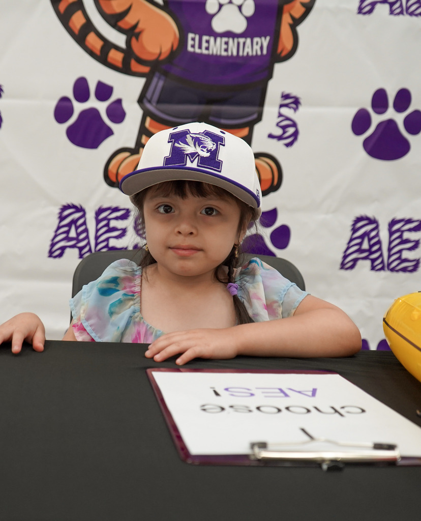 A little girl wearing a Minor High School hat sits at a table in front of an Adamsville Elementary School photo backdop and smiles.