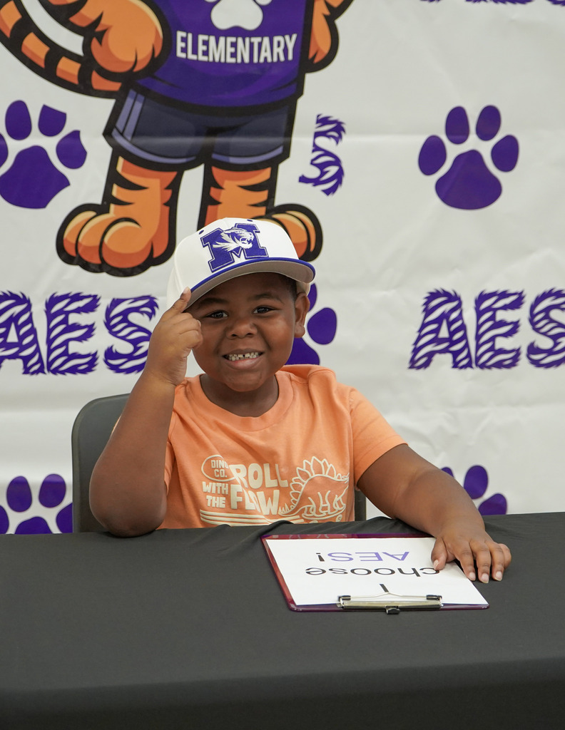 A little boy smiles while sitting at a table in front of an Adamsville Elementary School photo backdrop. He wears a Minor High School hat on his head and points toward it.