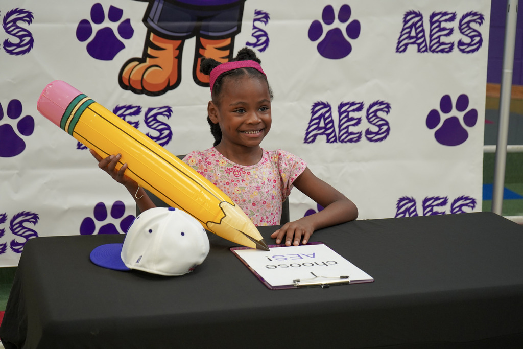 A little girl smiles while sitting at a table in front of an Adamsville Elementary School photo backdrop. She holds a large inflatable pencil prop in her hand.