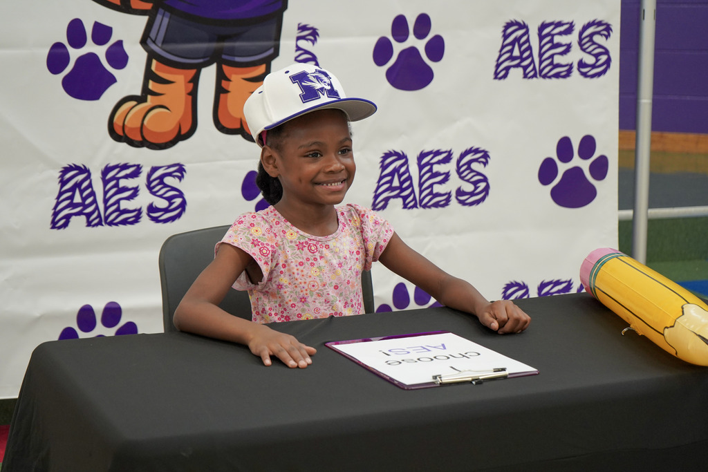 A little girl smiles for a photo while sitting at a table in front of an Adamsville Elementary School photo backdrop. She wears a Minor High School hat.
