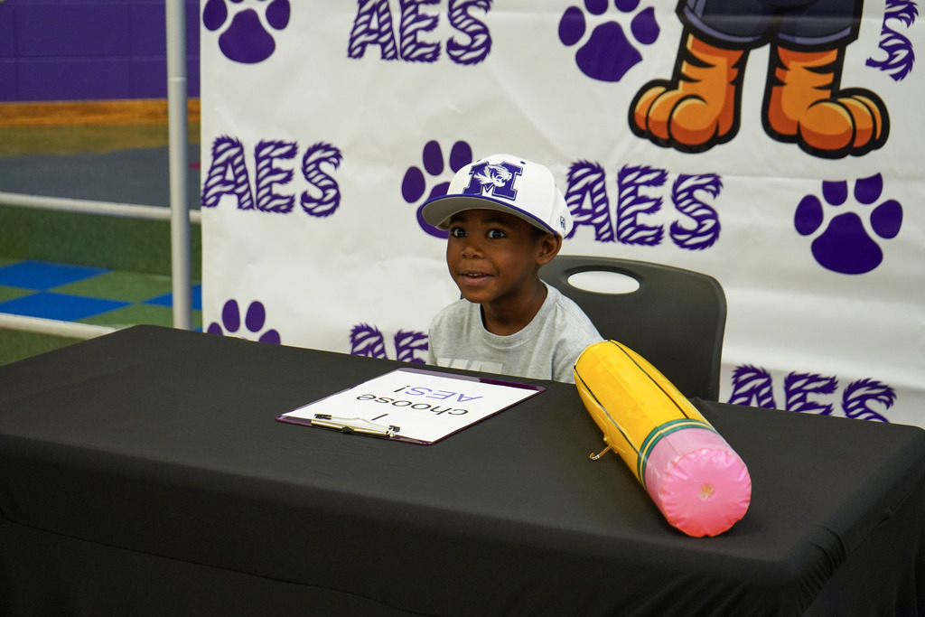 A little boy smiles while wearing a Minor High School hat and sitting at a table in front of an Adamsville Elementary School photo backdrop.