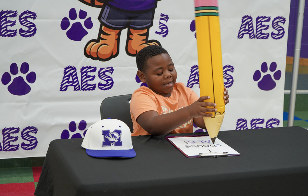 A little boy pretends to write with a large inflatable pencil while sitting at a table in front of a large Adamsville Elementary School photo backdrop.
