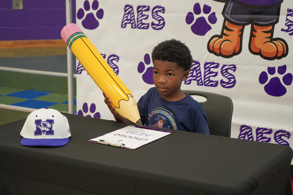 A little boy sits at a table in front of an Adamsville Elementary School photo backdrop and holds a large inflatable pencil prop in his hand.