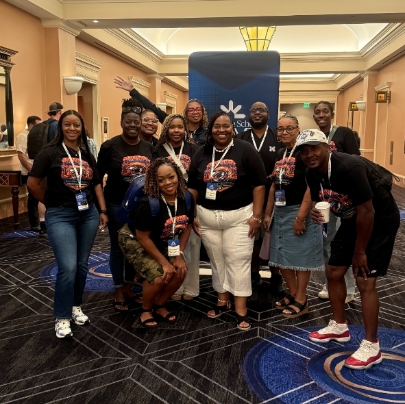 A wide shot of a large group of educators posing together in a wide, elegant hotel hallway. They are gathered around a vertical blue banner that reads "Innovative Schools Summit Exhibit Hall." To the far left, another banner displays "Thanks to our Sponsors" with corporate logos. The group is smiling for the camera, wearing matching black school graphic t-shirts.