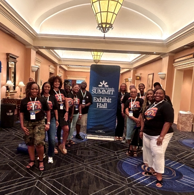 A group of eleven educators pose for a cheerful group photo in a hotel hallway in front of an "Innovative Schools Summit" banner. Most team members are wearing matching black t-shirts with a vibrant graphic design featuring a mascot and text. They are smiling, with one person waving in the back and another crouching slightly in the front row, showcasing a lively team dynamic.