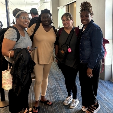 Four women stand side-by-side, smiling warmly for a photo in an airport terminal. From left to right, a woman in a grey t-shirt and glasses holds a black jacket; a woman wears a matching beige top and leggings with a backpack; a woman in a grey shirt and black pants wears patterned sneakers; and a woman in a denim jacket and black platform sandals poses with high-styled curly hair. A "Group 1" boarding sign is visible in the background.