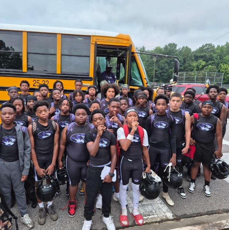 A youth football team in matching black and purple athletic gear posing together in front of a yellow school bus. Several players hold helmets, and the team is gathered on the asphalt under an overcast sky.