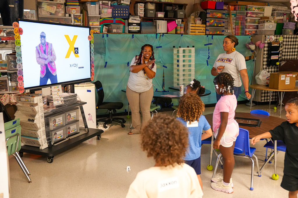 Students and teachers stand in a classroom and dance to an alphabet music video that's playing on a smart board.