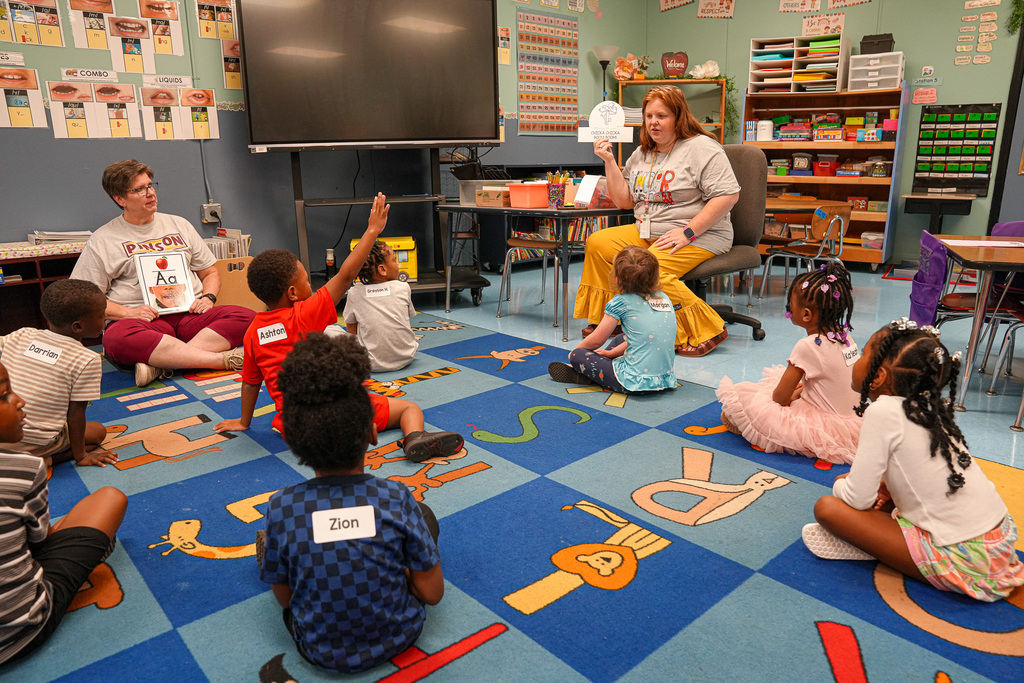 A teacher holds up a piece of paper for students to look at while the children sit on a colorful rug on the floor of the classroom.