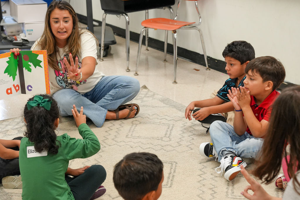 A teacher holds up the number three while reading a book to students as they all sit on the floor of a classroom.