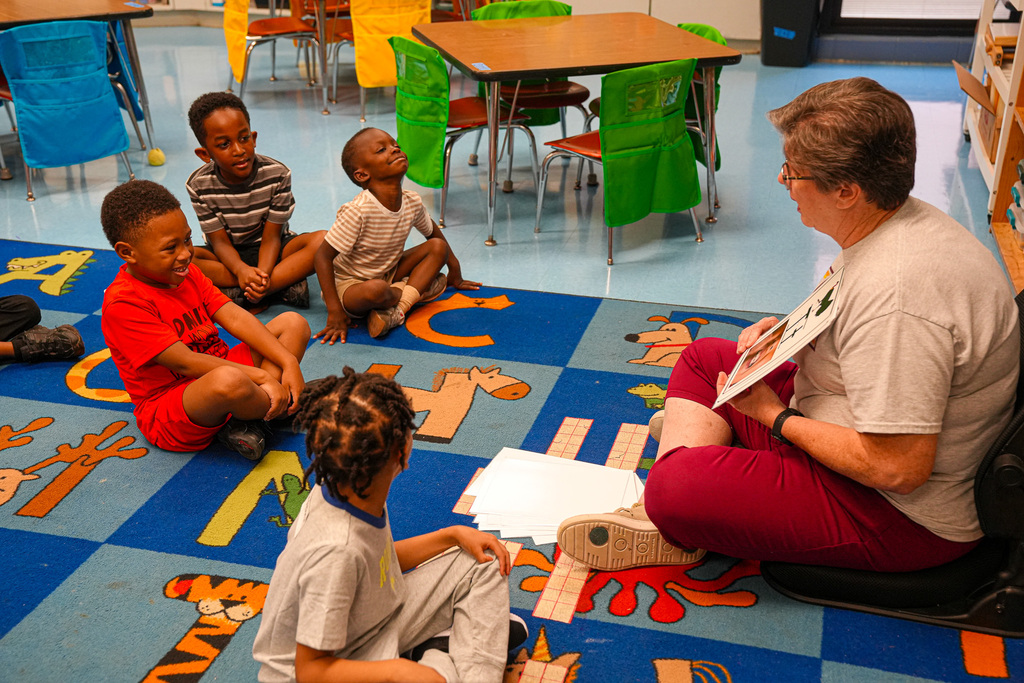 A teacher sits on the floor of a classroom with students. The teacher holds up large cards with letters on them for the students to read.