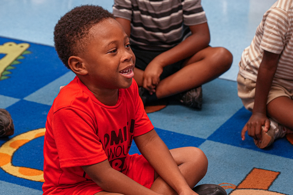 A little boy smiles while sitting on a colorful rug on the floor of a classroom.