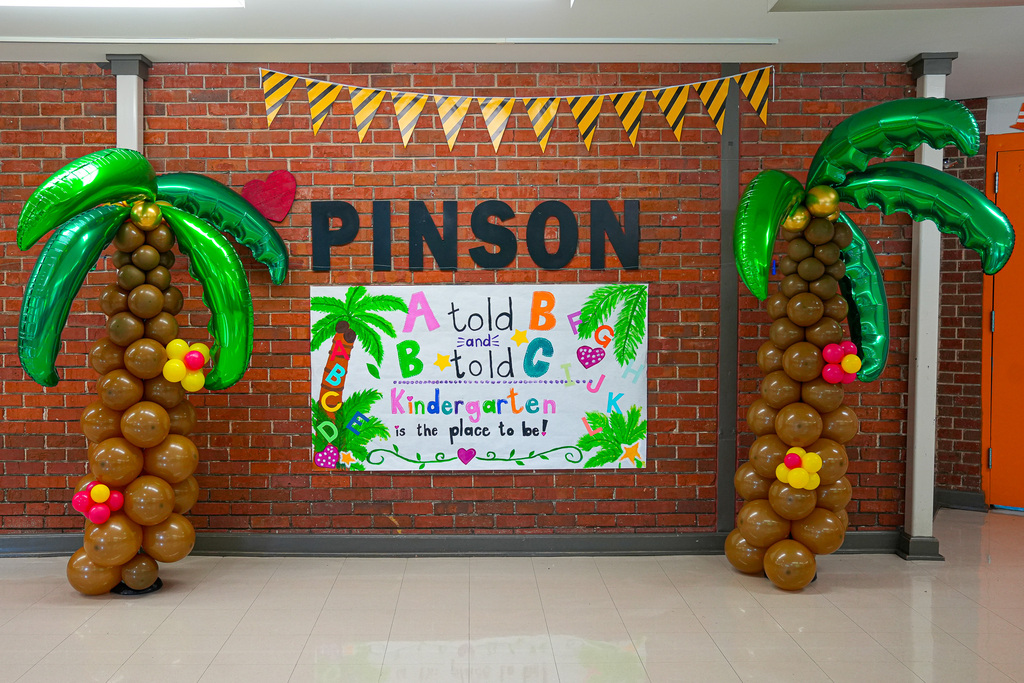 A sign on the wall of Pinson Elementary School says "A told B and B told C, Kindergarten is the place to be." Beside the sign are large balloon decorations made to look like palm trees.