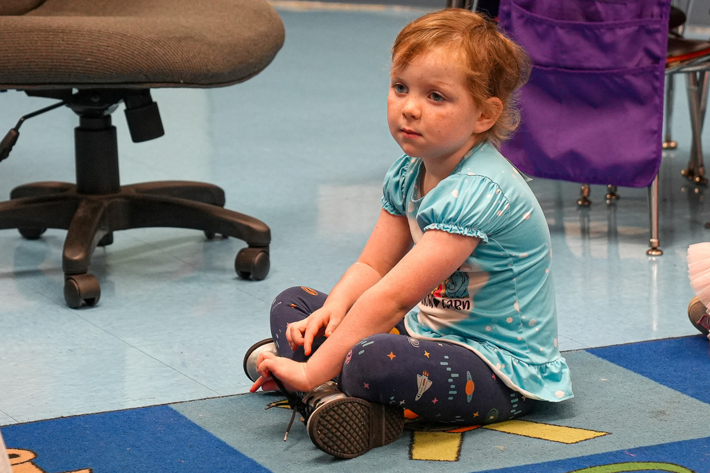 A student sits on a colorful rug on the floor of a classroom and listens to the teacher.