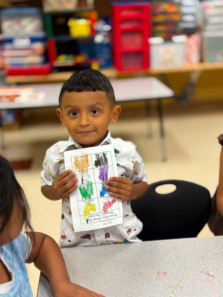 A little boy smiles and holds up a piece of paper that he colored on with crayons.