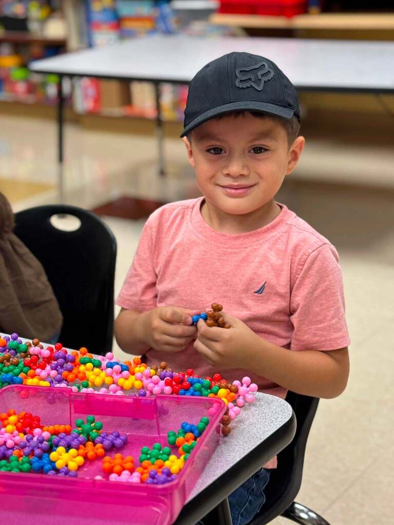 A child smiles while holding a toy in his hand. He sits at a table inside a classroom.