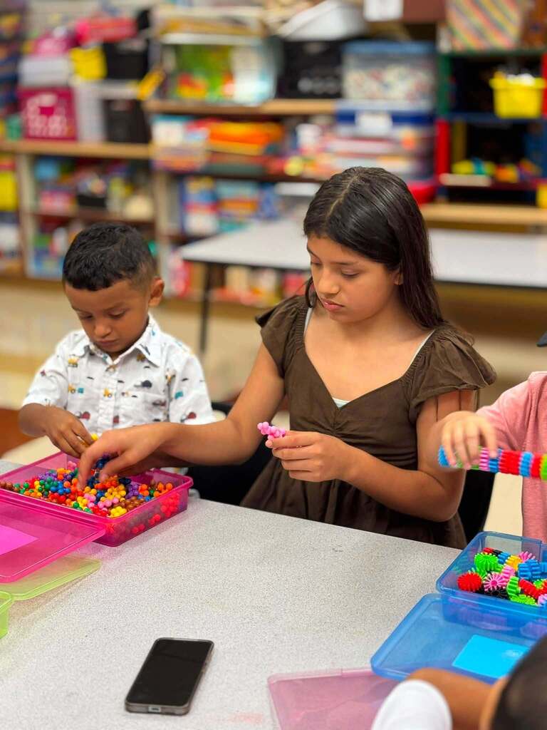 An older student helps a younger student build small structures while they sit at a table inside a classroom.