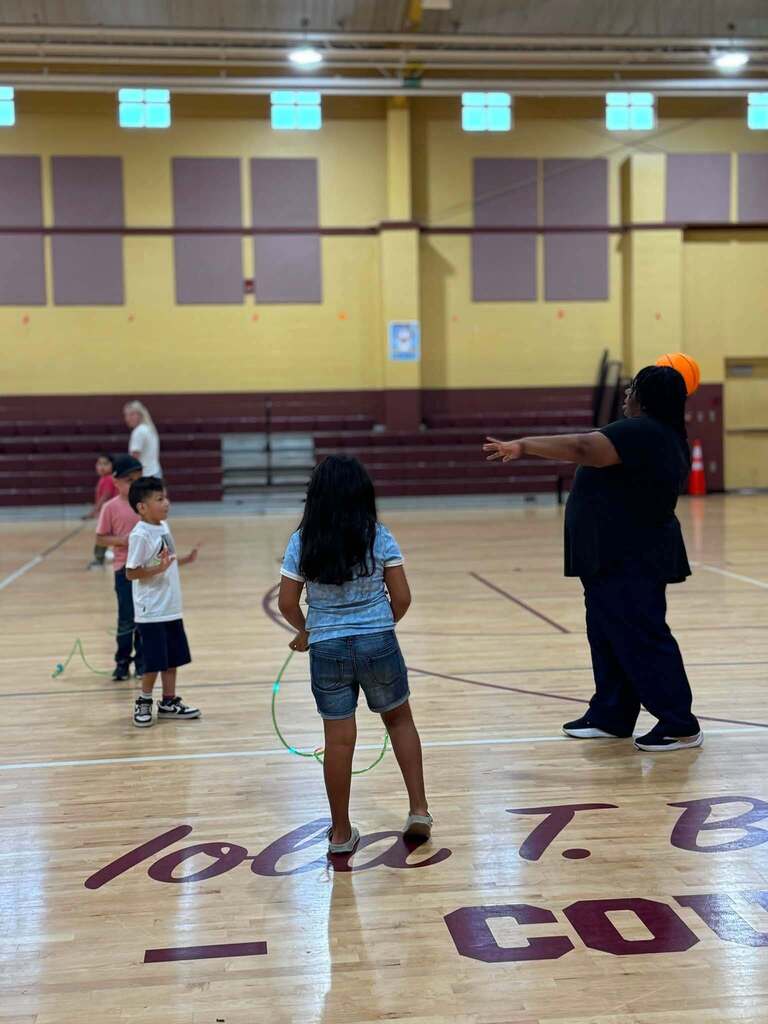 A teacher throws a basketball while students play around her inside a school gym.