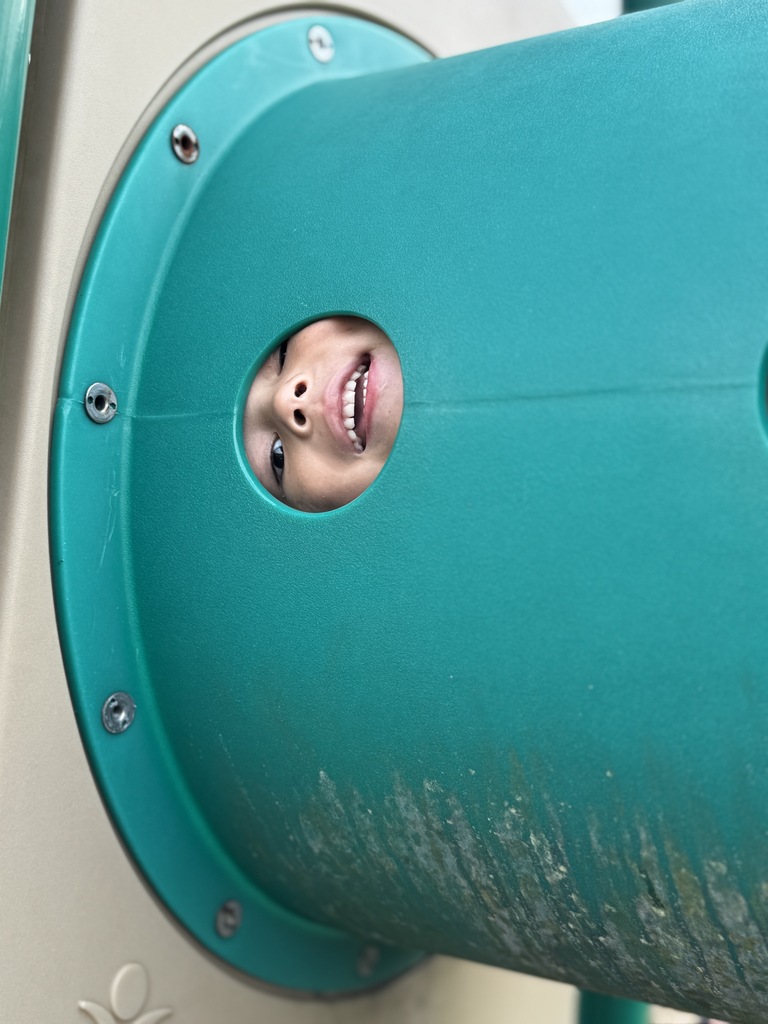 A child smiles while looking through the hole of a playground slide.