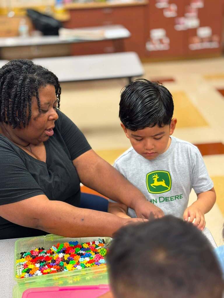 A teacher and a student stand and sit at a table inside a classroom. The teacher helps a student as they build with legos.