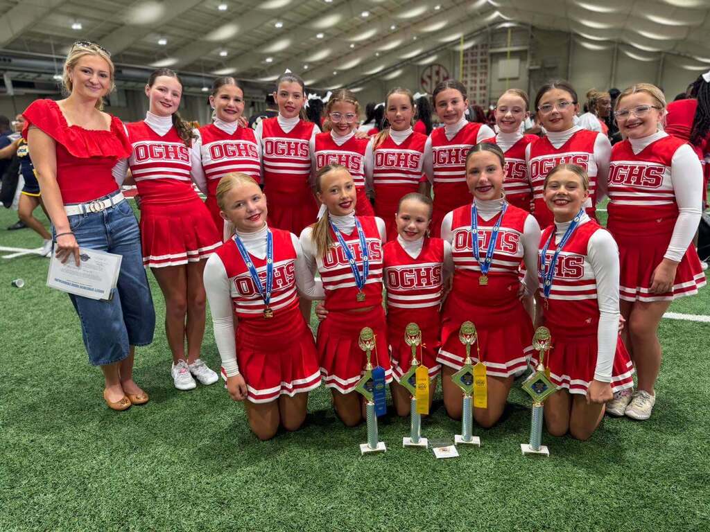 Oak Grove High School cheerleaders pose for a photo inside a football training facility. They wear their cheer uniforms and pose behind trophies.