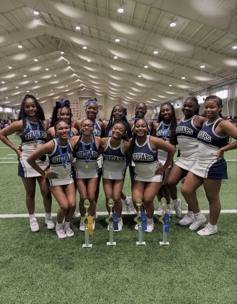 Clay-Chalkville High School cheerleaders stand together for a photo inside a practice facility. They wear matching cheer uniforms and stand behind several trophies.