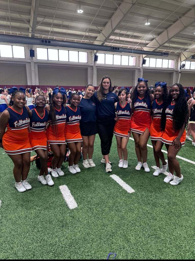 Fultondale High School cheerleaders pose for a photo inside a football training facility. They wear their uniforms and stand on the turf.