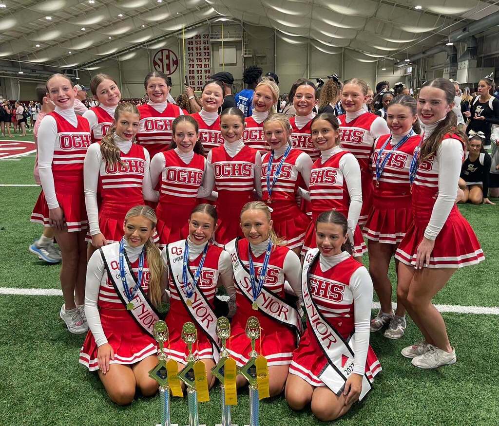 Oak Grove High School cheerleaders pose for a photo inside a football training facility. They wear their cheer uniforms and stand and kneel behind several trophies.