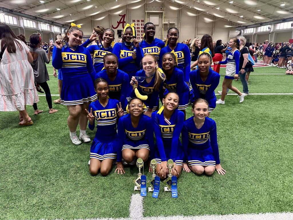 JCIB cheerleaders pose for a photo inside a football training facility. They smile and pose behind several trophies.