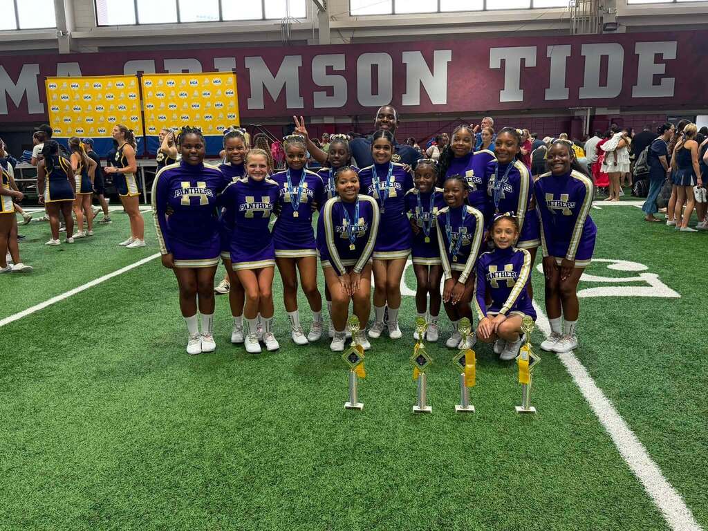 Hueytown High School cheerleaders pose for a photo on a practice field. They wear matching purple cheer uniforms and pose behind several trophies.