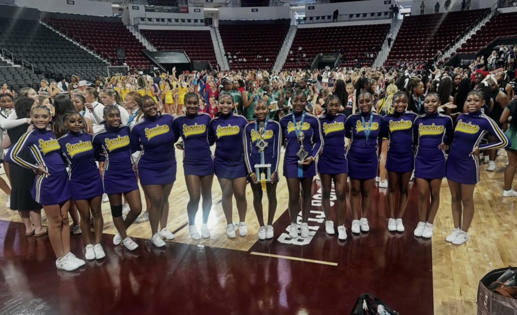Pleasant Grove High School cheerleaders pose for a photo inside a gym. They wear matching purple cheer uniforms and two of the girls hold trophies.