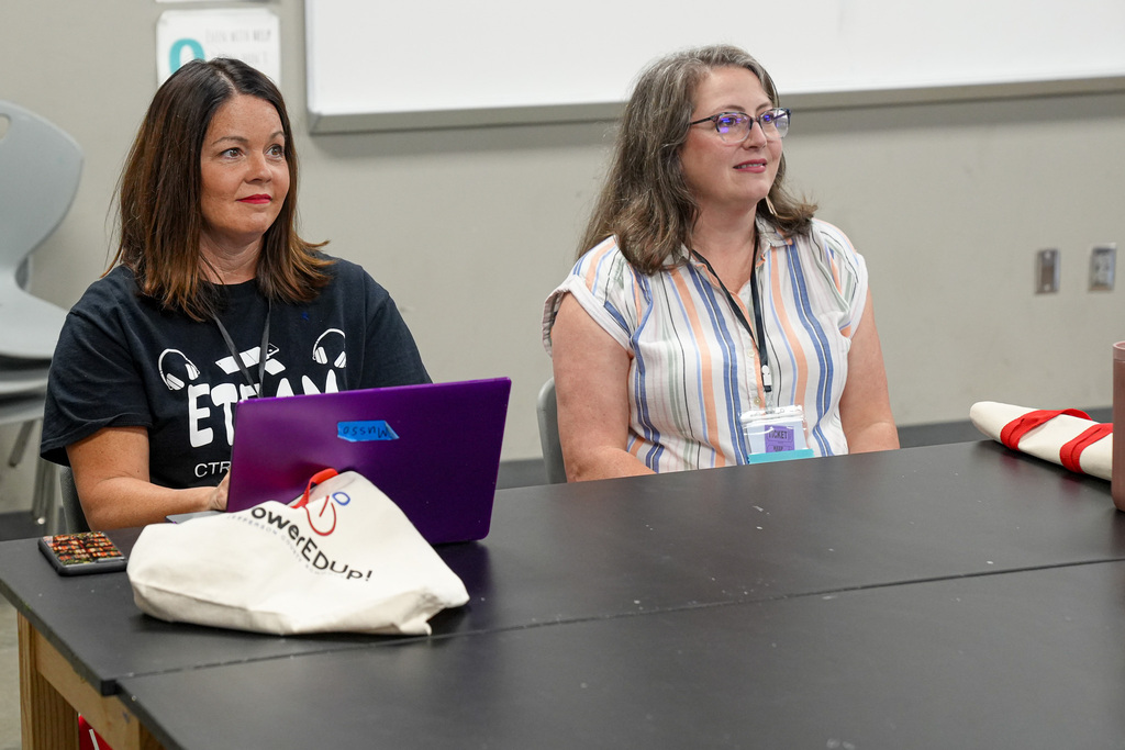 Two women smile while sitting at a table inside a classroom.