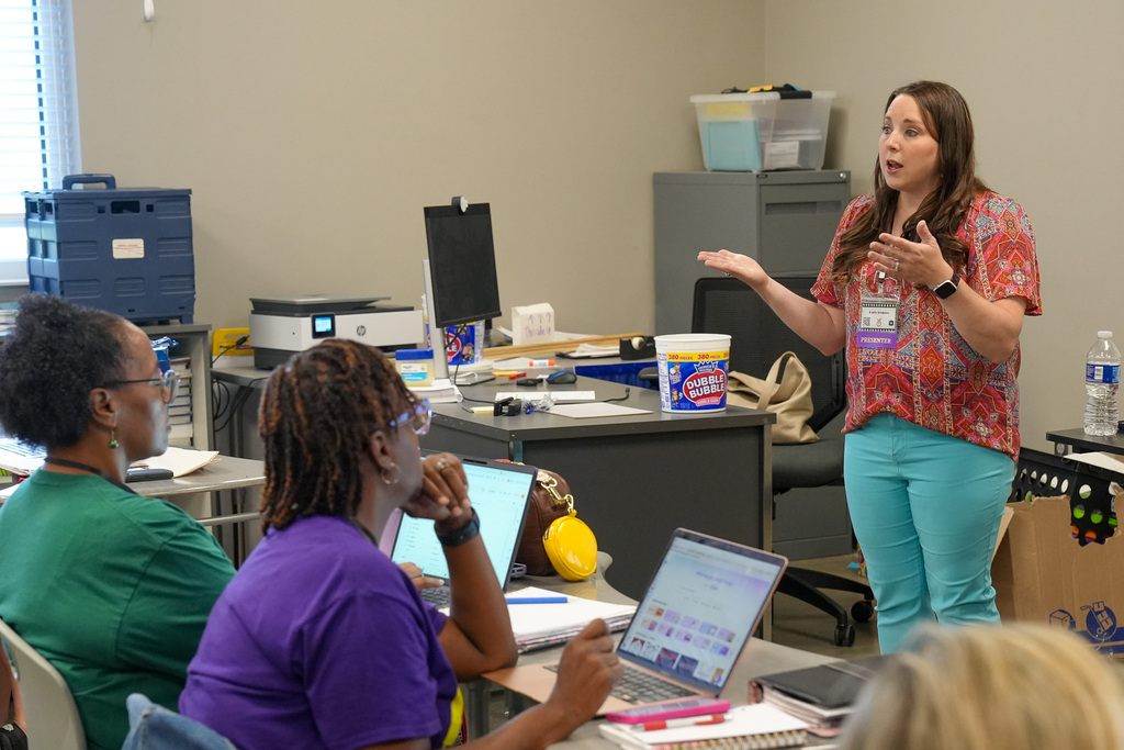 A woman stands in front of a classroom of educators and speaks to them as they sit at tables and take notes.