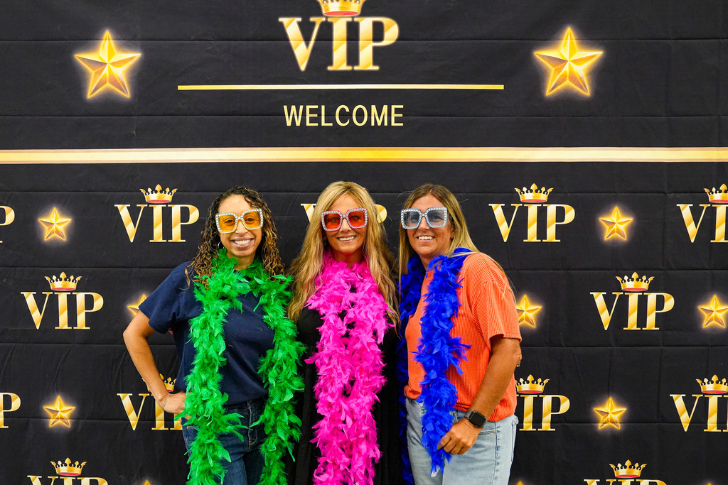 Three women stand together for a photo. They smile and pose in front of a photo backdrop with stars and "VIP" written on it. They wear sparkly sunglasses and feather boas.