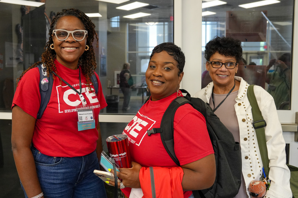 Three women smile for a photo while standing inside a room. Two of the women wear matching CTE shirts.