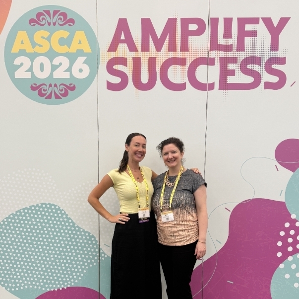 Two smiling women stand side-by-side posing in front of a large event backdrop. The backdrop features the words "ASCA 2026" inside a circular logo on the left and the phrase "AMPLIFY SUCCESS" in large, bold purple letters on the right, surrounded by colorful teal and magenta abstract shapes. The woman on the left is wearing a yellow top, a long black skirt, sneakers, and a yellow event lanyard. The woman on the right has her arm around her colleague's shoulder and is wearing a textured grey-to-peach gradient t-shirt, black pants, sandals, and a yellow event lanyard.